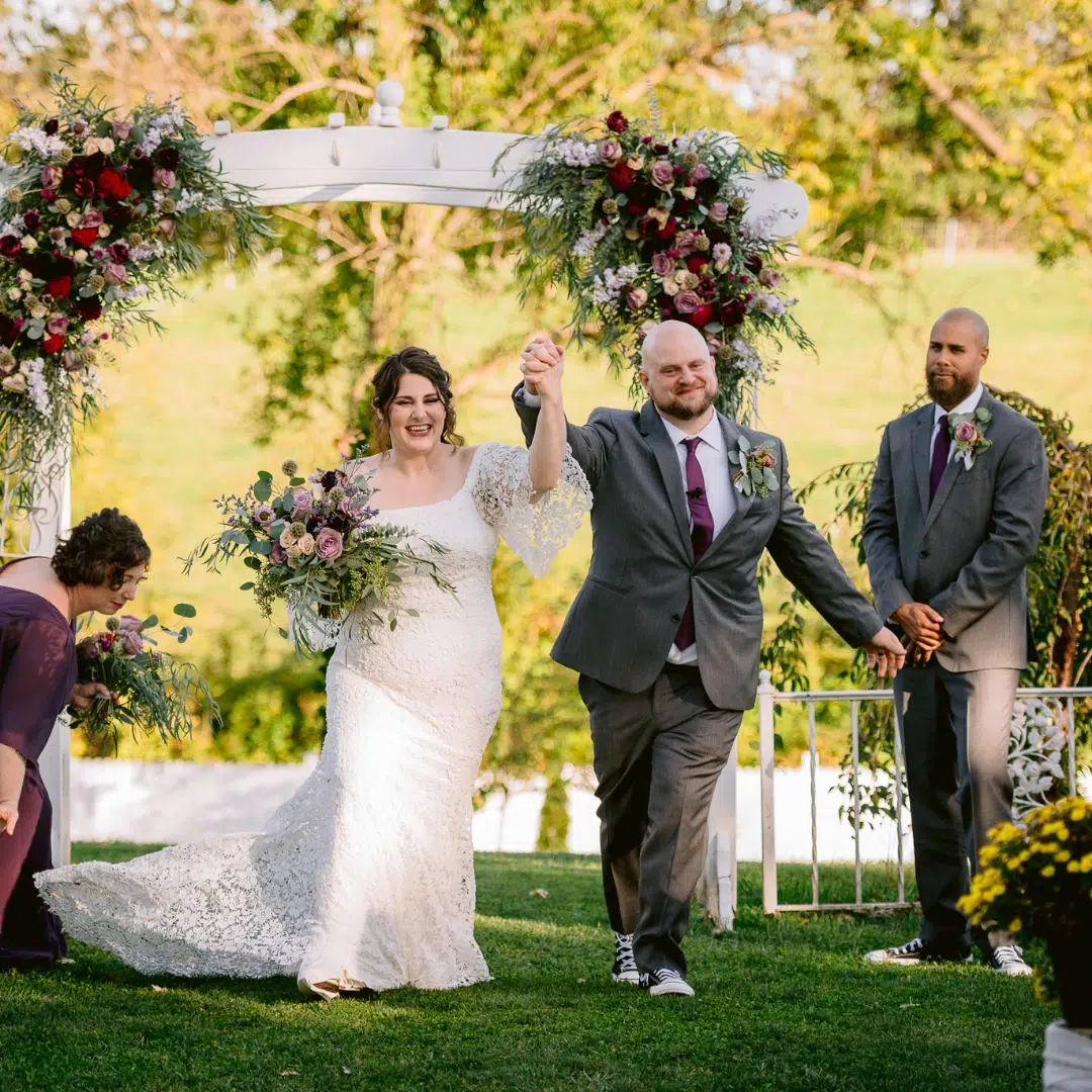 Photo of a groom and a bride holding a bouquet of purple flowers in a park with a white arch behind them.