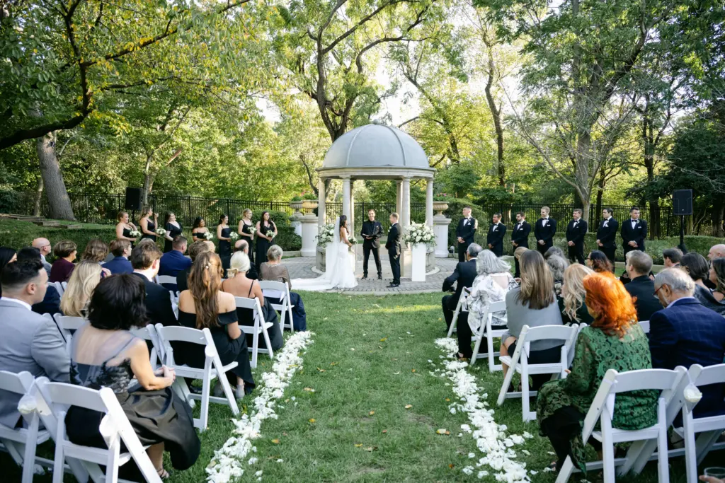 Photo of a wedding ceremony in a garden with seated guests and white flower decorations.