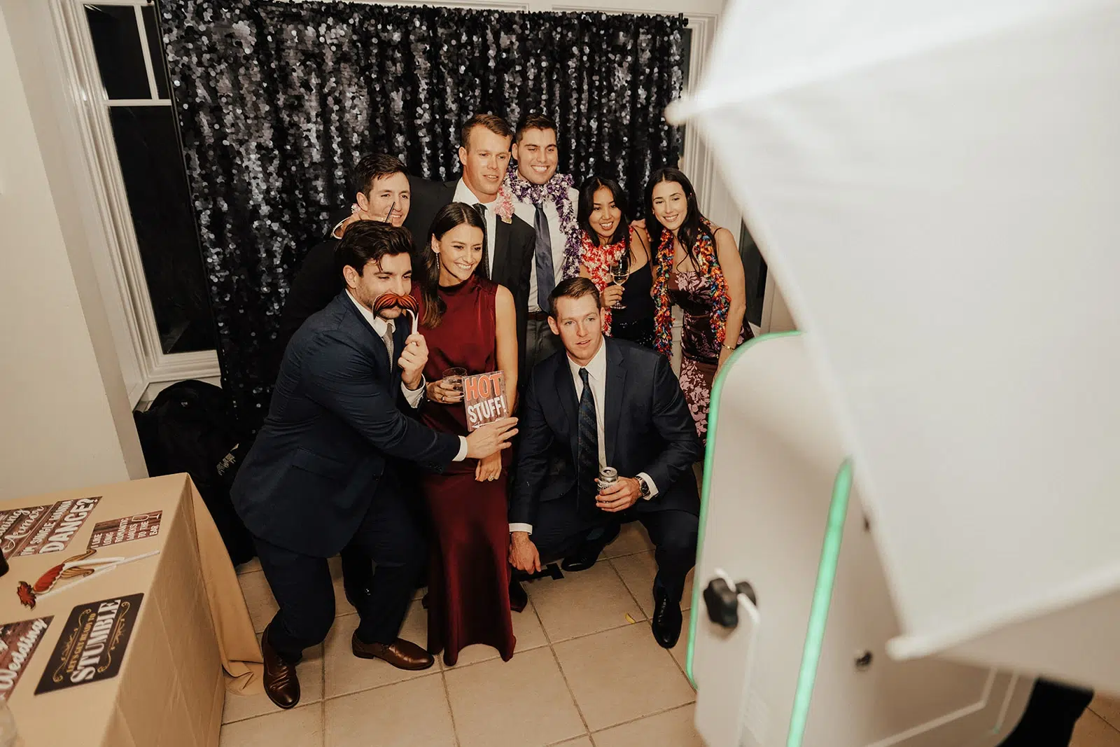 Photo of wedding guests taking pictures at a photo booth with signs and a sparkly backdrop.
