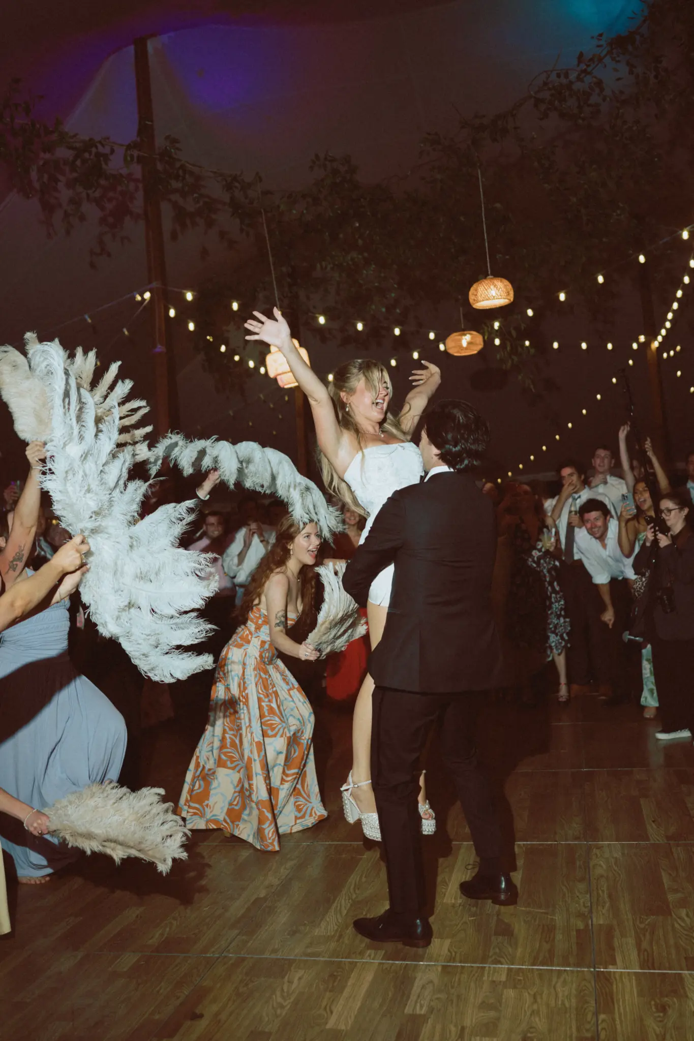 Photo of a groom carrying his bride on the dance floor while their guests are holding out large feathered fans.