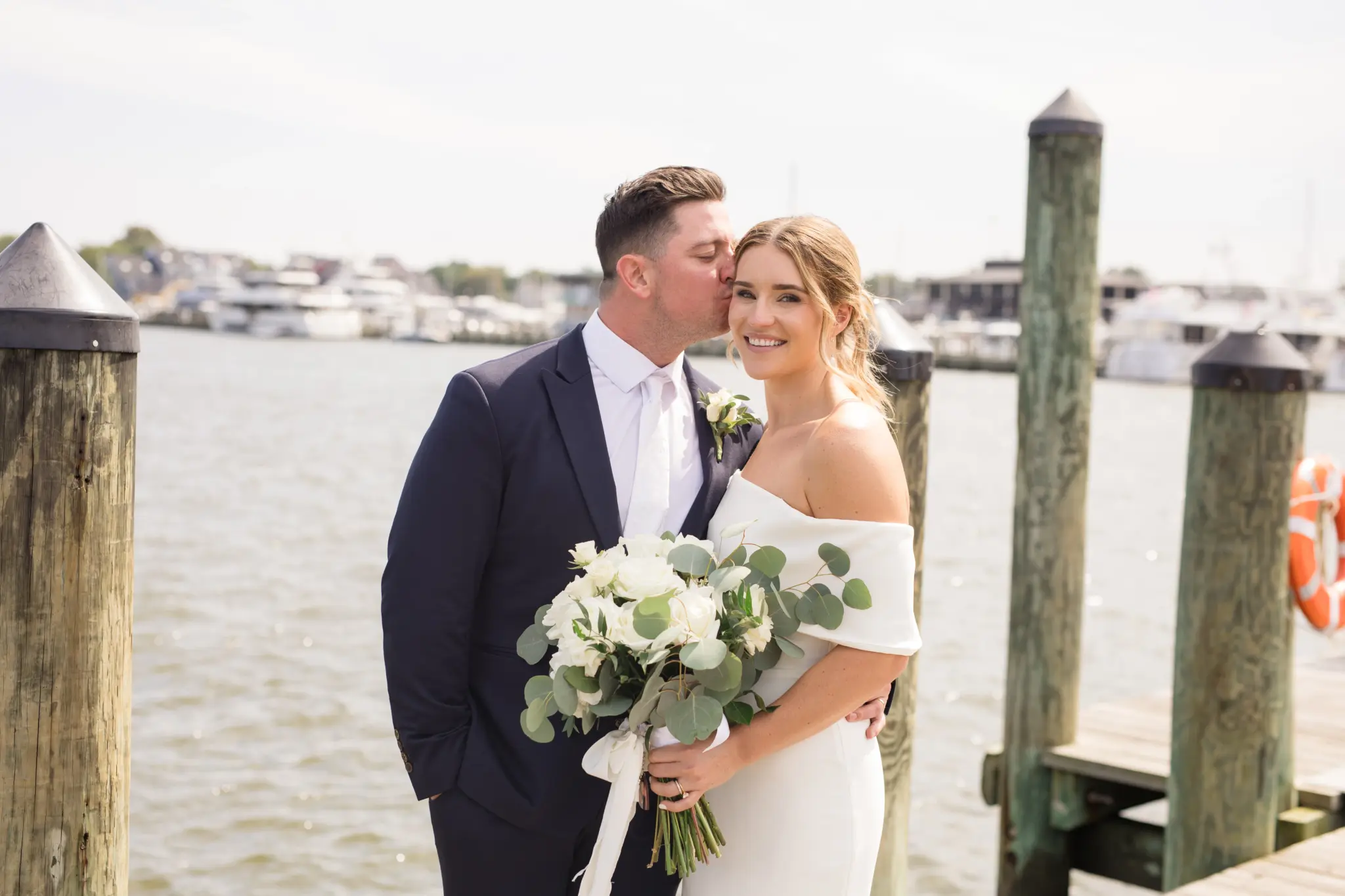 Photo of a groom kissing a bride who is holding a bouquet of white roses on a dock.