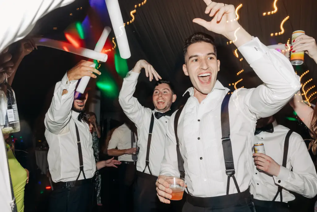 Photo of a groom and his groomsmen dancing at a wedding with drinks in their hands