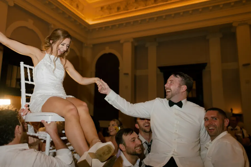 photo-of-a-groom-holding-his-bride's-hand-while-she's-seated-on-a-chair-carried-by-the-crowd