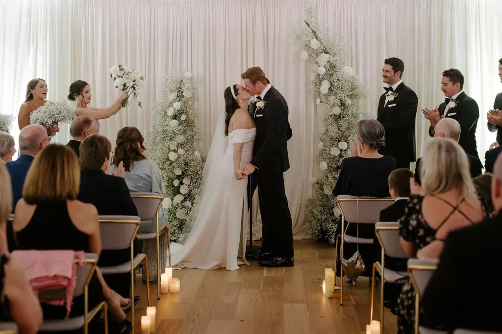 photo of a bride and groom kissing at a wedding ceremony while their guests are seated and their bridesmaids and groomsmen are clapping