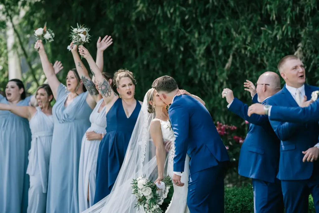 photo-of-a-bride-and-groom-kissing-at-their-wedding-ceremony-while-the-bridesmaids-in-blue-dresses-and-groomsmen-in-blue-suits-cheer-for-them