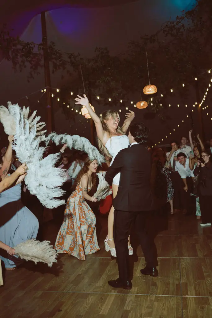 photo of a groom carrying a bride on the dance floor with her hands up in the air and while the guests hold big feather fans