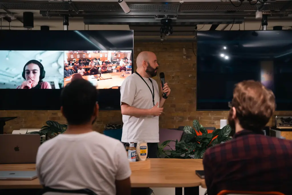 photo of a man speaking into a microphone with a video meeting on a screen at a hybrid event